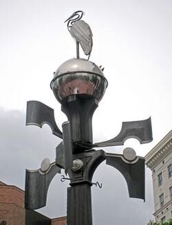 Photograph of the top of a lamp post-like apparatus. A bird figure sits atop a globe on the top, and four appendages jut out at ninety-degree angles from each other on the post.