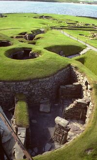 Various stone-lined depressions have been cut into a sward-covered area near a body of water. Some depressions are small and circular. A larger one in the foreground is semi-circular and contains various arranged piles of stones.