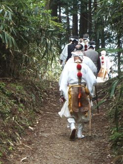 Monks with cropped hair and walking staves hiking up a steep mountain trail in white robes.