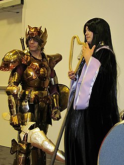Two people in costume, a brown-haired man wearing gold armor and a woman with long black hair wearing a dress.
