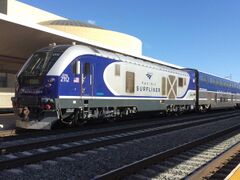 A diesel locomotive with navy blue curved shapes on the front and rear with white accents, a black cab area, and Amtrak California logos on the front and sides