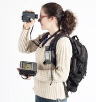 A Woman with a backpack holding a laser rangefinder, a handheld GPS and a Tablet computer.