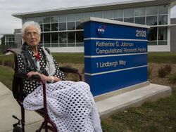 Johnson seated on a bench beside a sign in front of the Katherine G. Johnson Computational Research Facility.