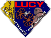 A diamond-shaped crest houses artworks of the Lucy fossil at left, the Lucy spacecraft at center, and an artist's impression of a Jupiter trojan. The word "Lucy" is written in a large, bold red font at the top right corner, while the words "First to the Trojans" and "SWRI · NASA · LM" are written in a smaller white font across the bottom edges of the diamond-shaped crest.