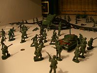 Two teams of green soldiers, each consisting of a variety poses, one having a tank, and another having a truck, are separated by a black wire on a kitchen countertop. Behind them is a large black wine bottle and a brown basket.