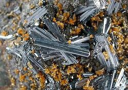 A close view of a rock crusted with groups of glassy, lustrous, silvery-blue hutchinsonite, in tight clusters of loosely aligned needle-like crystals, among smaller clusters of tiny orange-brown crystals