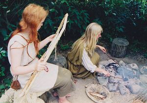 Two women in ancient costumes, one spinning a loom and the other making a fire