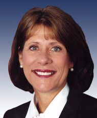 A brown-haired woman in her early fifties wearing a white blouse, black jacket, and pearl earrings