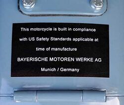 Black placard mounted on a light blue fuel tank, which reads "This motorcycle is built in compliance with US safety standards applicable at the time of manufacture BAYERISCHEN MOTOREN WERKE AG Munich / Germany"