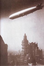 A sepia photograph of the airship from below. It is flying from right to left above a city. There is a tall ornate building just below the airship and the silhouette of more masonry in the bottom left foreground. All five engine nacelles are visible, and the two nearest and the control room are in sunlight. There are swastikas on both the vertical fins.