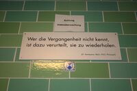 A green brick wall with a white sign reading "Wer die Vergangenheit nicht kennt, / ist dazu verurteilt, sie zu wiederholden. / (G. Santayana 1863–1953, Philosoph)