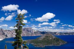 Wizard Island is shown in Crater Lake, with clouds in the sky above. The caldera rim appears to the left.