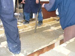 A handheld concrete vibrator consolidates fresh concrete in wooden formwork for a concrete beam.