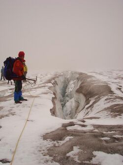 A linear crack in a glacier with a person equipped with climbing gear standing to the left.