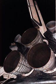 Three bell-shaped rocket engine nozzles projecting from the aft structure of a Space Shuttle orbiter. The cluster is arranged triangularly, with one engine at the top and two below. Two smaller nozzles are visible to the left and right of the top engine, and the orbiter's tail fin projects upwards toward the top of the image. In the background is the night sky and items of purging equipment.