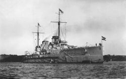 A large gray ship in port. The two funnels in the center of the ship emit clouds of smoke.