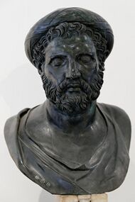 Bronze bust of a man with a short, curly beard wearing a tainia, which resembles a turban. Short curls hang out from underneath the tainia. The face is much broader than the other busts and the neck much fatter. The brow ridges are very prominent.