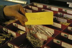 A person's hand is shown holding a transparent plastic bag of bolts (machine screws) together with a yellow card. The card has printed on it: "ITEM #0014 BIN: A14 NSN:5306-00-151-1419 P/N: NOMEN: BOLT QTY LEFT: 50".