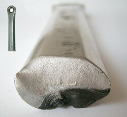 A roughly ovoid metal cylinder, viewed end-on. The bottom-right portion of the metal's end surface is dark and slightly disfigured, whereas the rest is a much lighter colour and not disfigured.