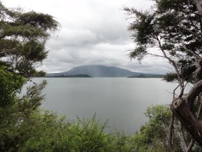 Image showing a still Lake Rotomahana on an overcast day framed by trees. Mount Tarawera is visible in the background