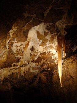A long stalactite hangs from the ceiling of a cave passage, with some smaller calcite stalactites nearby.