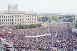 A crowd of thousands stands in a plaza. Many carry large banners.