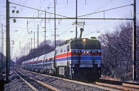 A passenger train led by an electric locomotive with black roof, light grey sides, and horizontal red and blue stripes separated by thin white stripes. The passenger cars are stainless steel with stripes matching the locomotive.
