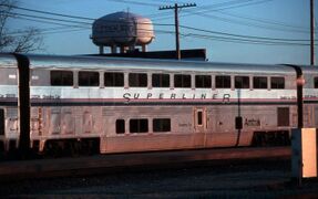 A stainless steel bilevel passenger rail car. A blue stripe and two thinner red stripes are on the side between the two levels of windows.