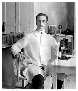 Black and white photo of Charlos Chagas, in his lab coat, sitting next to his microscope and surrounded by flasks and jars