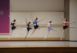 Five dancers leap on a stage. Each wears a number on her leotard.
