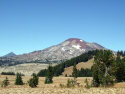 South Sister, which features patches of snow and ice, can be seen above a plain. Forest is visible in the foreground.