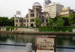 Atomic bombing memorial in Hiroshima. The remains a building with the skeletal structure of a dome at the top sits among modern Japanese buildings and green space.