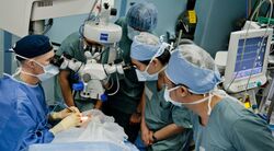 A surgical team is gathered around the patient in an operating theatre. the surgeon is looking through a surgical microscope suspended above the patient's eye.