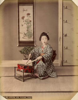 A woman wearing a kimono and traditional hairstyle kneels on a tatami mat, playing a drum in front of her with two sticks.