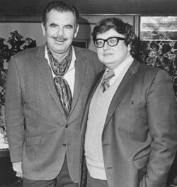 A black and white photograph of two men in suits. The man on the right is wearing glasses.
