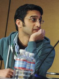 A man wearing a teal hoodie looking to the left. He is seated at a table behind a microphone and water bottles.