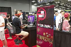 A man with red hair, a black shirt, and cargo shorts plays a demo of Hotline Miami 2 with a video game controller. Several other people are in the background, and promotional material for the game sits next to him.