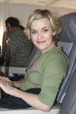 A woman sits in an airplane seat, smiling towards the camera. She has short blond hair.