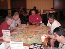 Five men sit at a table, poring over a map. Dice and counters lie atop the map, some counters are stacked. The map is subdivided into small hexagons of identical size.
