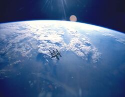 A cluster of modules and feathery solar arrays floats in the middle distance before an image of the Earth and the blackness of space above its horizon. Sunrays project from the top centre of the image.