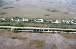 A color photograph taken from the air showing the Everglades bisected by a highway; at the bottom is a sawgrass field flooded with water bordered by a full canal; at the top are some homes and a dry sawgrass field