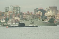 A submarine sitting in calm water, in front of a large warship, and with numerous tall buildings in the background. White-uniformed personnel are standing on the decks of both vessels.