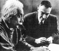 Einstein writing at a desk. Oppenheimer sits beside him, looking on.