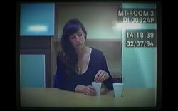 Old computer footage of a 37-year-old woman with long brown hair in a red jacket, sitting at a police interview desk. She is holding her hands together in front of her, and is looking to the right of the camera.