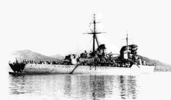 Water-level rear quarter view of a large grey warship at anchor. One gun turret, the main mast and both funnels are prominent.