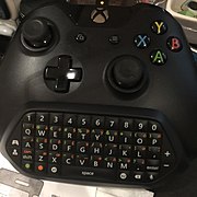 Microsoft Chatpad attached to an Xbox One Wireless Controller; the Chatpad is a black plastic keyboard that fits between the two handles on the bottom of the Xbox controller, providing dedicated hardware keys for alphanumeric text input, arranged in a QWERTY layout with a number row at the top and a long spacebar at the bottom. Flanking the QWERTY keys are four keys that provide the same functionality as the Stereo Headset Adapter. On the bottom row are two modifier keys (green and orange) for special characters, a chat microphone mute button, and programmable keys marked X1 and X2, immediately flanking the spacebar.