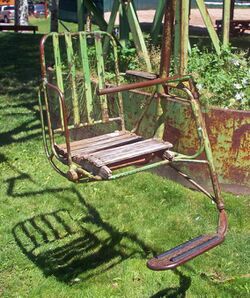 A metal chair, painted green, with footrest and wooden seat suspended above a grassy area by a metal pole on the right side