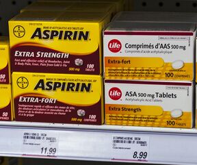 Four boxes of medication on a store shelf above price tags. The two on the left are yellow with "Aspirin" in bold black type and explanatory text in English on the top box and French on the bottom. The two on the right are slightly smaller and white with the word "Life" in the corner inside a red circle. The text, in French on top and English below, describes the medication as "acetylsalicylic acid tablets"