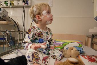 A child sits on a hospital bed in pyjamas with soft toys. Along with other measurement devices, the child has electrodes taped to their scalp and face.