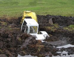 Photograph of a backhoe that is over half submerged in a large hole that it dug in a peat bog before falling in.